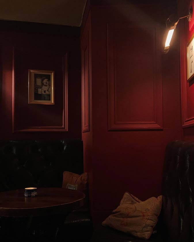 Dark corner of a bordeaux colored bar. Lived in cushions and gold picture frames. The only light sources are a table candle and a dim wall sconce.