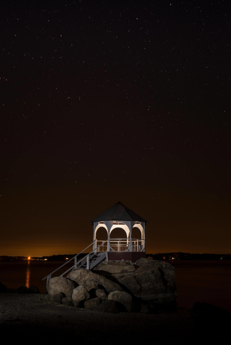 A gazebo, built on ice age rocks, sits on the shore; a distant town is in the background while stars are prominent in the sky.