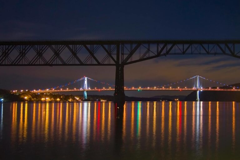 The Walkway over the Hudson and the Mid-Hudson Bridge are shown in a nighttime scene, with their lights reflected in the water